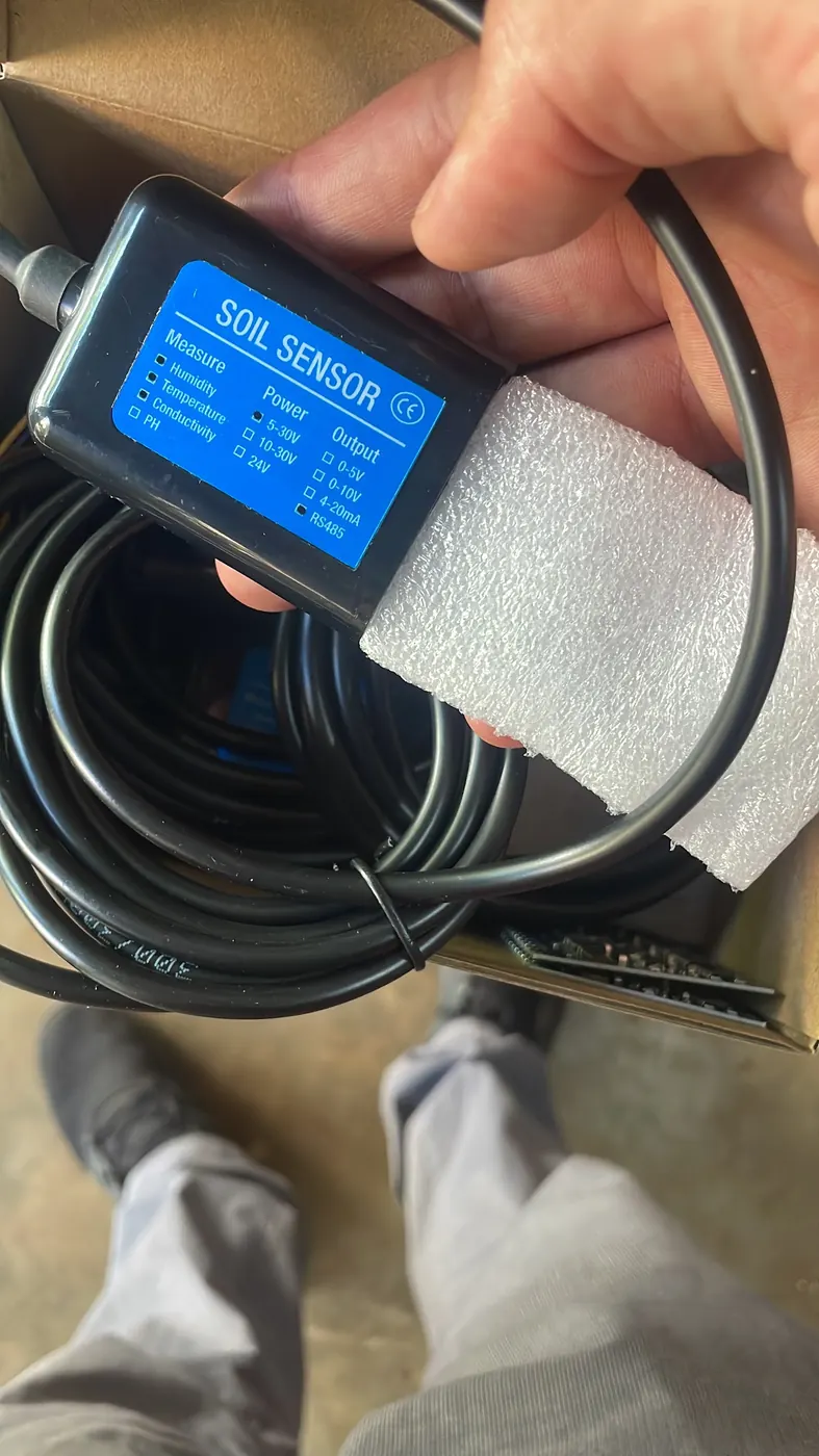Hand holding a black soil sensor with blue label and coiled cable above an open cardboard box.